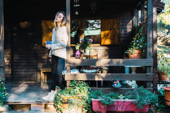 Junge sinnliche Frau mit Morgenkaffee im Becher, während sie auf der Veranda des Holzhauses posiert. — Stockfoto