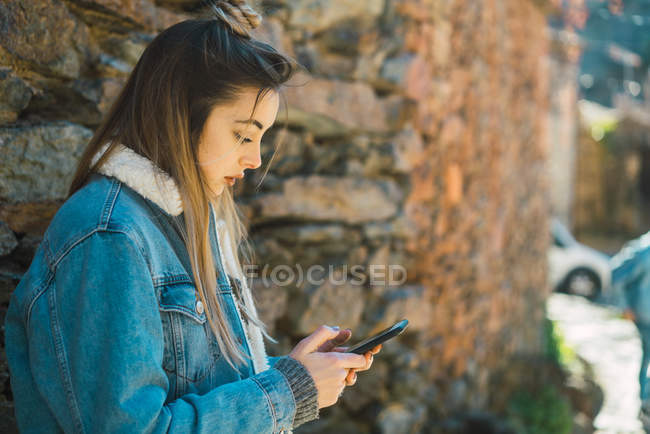 Вид збоку на стильну дівчину з використанням смартфона на фоні кам'яної стіни — стокове фото