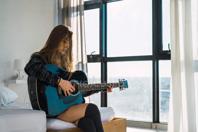 Молода жінка сидить на ліжку і грає на гітарі вдома — стокове фото