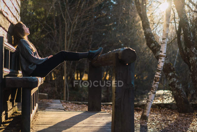 Side view of casual woman sitting on wooden bench outside of rural house in autumn sunlight. — Stock Photo