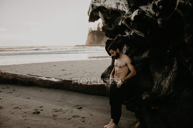 Vista lateral del hombre sin camisa apoyado en el tronco caído en la playa - foto de stock