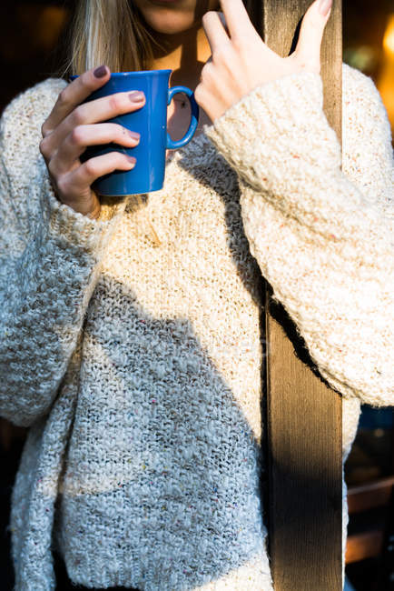 Crop of woman in cozy sweater embracing wooden pillar on porch and enjoying coffee. — Stock Photo