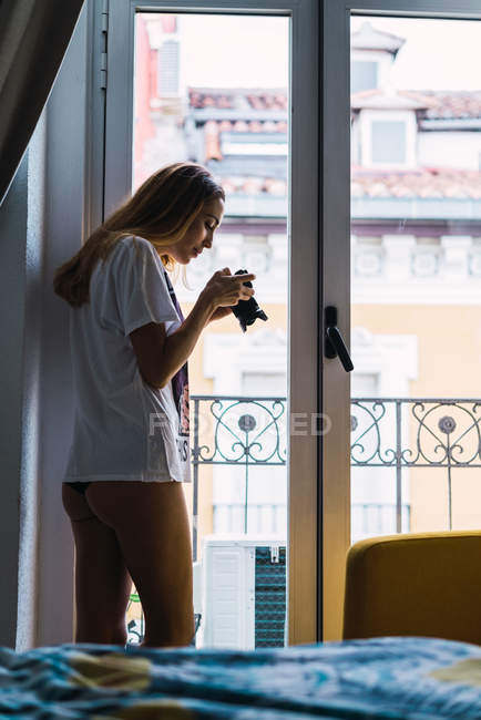 Vue latérale de la photographe femme regardant à travers la caméra dans la fenêtre à la maison . — Photo de stock