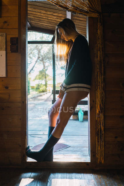 Vista lateral da jovem mulher em meias e suéter em pé na porta da casa rural . — Fotografia de Stock