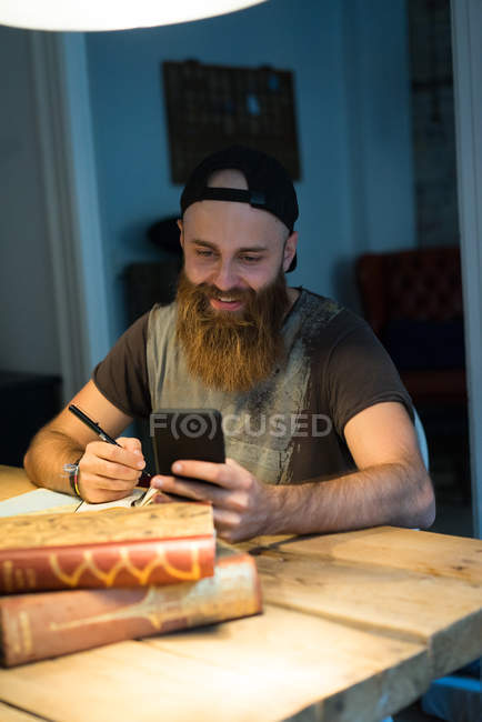 Porträt eines bärtigen Mannes, der Telefon benutzt und am Tisch schreibt — Stockfoto