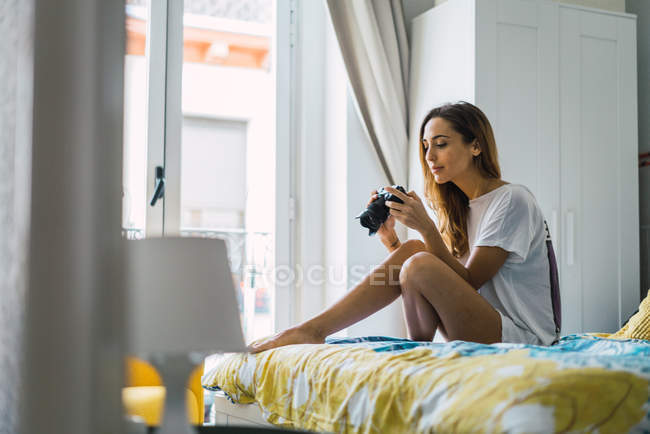 Retrato de mujer casual sentada en la cama y mirando fotos en la cámara en casa - foto de stock