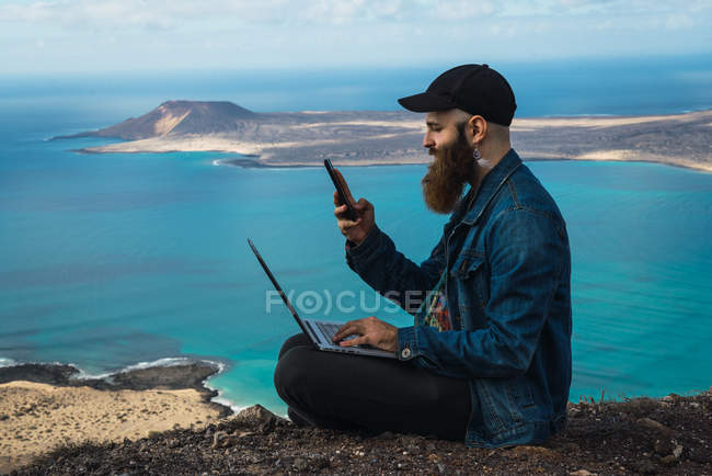 Seitenansicht eines Mannes, der mit einem Laptop auf Knien auf einer Klippe sitzt und an sonnigen Tagen im Smartphone surft — Stockfoto