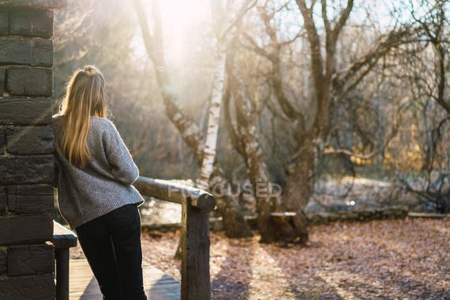 Back view of woman in sweater leaning on wall of wooden rural house at autumnal landscape. — Stock Photo