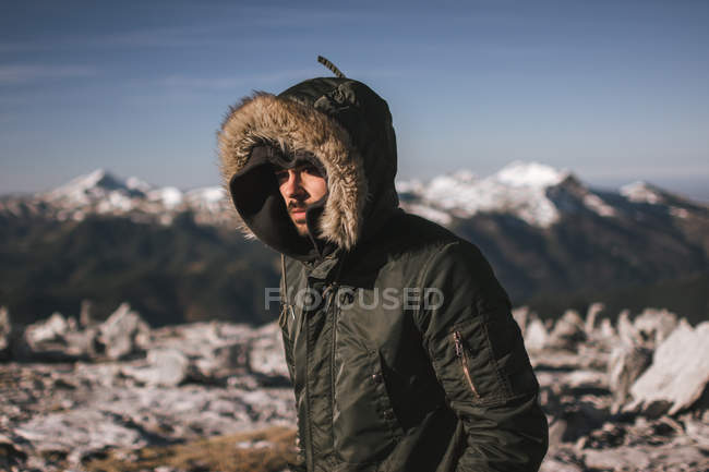 Porträt eines Mannes in warmem Mantel mit Kapuze, der im Sonnenlicht vor dem Hintergrund schneebedeckter Berge steht. — Stockfoto
