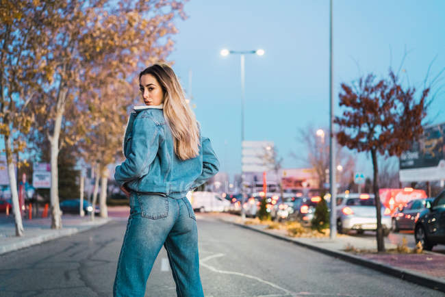 Вид спереду молодої жінки в джинсовому одязі позує на вулиці і дивиться через плече на камеру — стокове фото