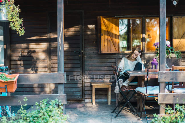 Donna che legge libro in poltrona al portico chalet — Foto stock
