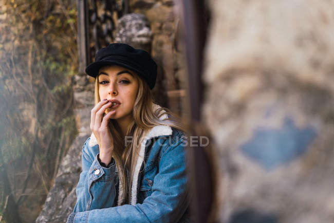 Portrait of young woman in cap and jacket touching lips sensually and looking at camera — Stock Photo