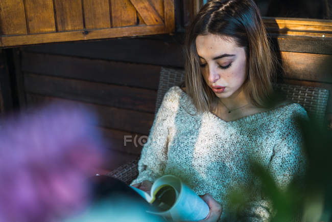 Vista de ángulo bajo de chica rubia que se enfría en sillón en el porche y libro de lectura con concentración . - foto de stock