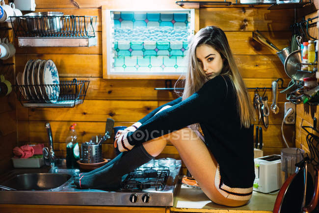 Vista lateral de la mujer joven en suéter y calcetines sentados en el mostrador en la pequeña cocina y posando sensualmente . - foto de stock