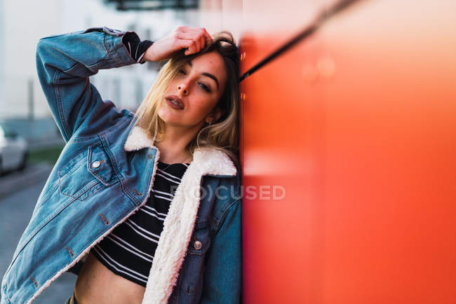 Selbstbewusstes blondes Mädchen in Jeans lehnt an roter Wand und schaut in die Kamera, während sie den Kopf berührt. — Stockfoto