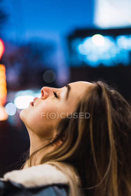 Vue latérale de blonde sensuelle posant avec les yeux fermés sur fond de lumières de ville floues — Photo de stock