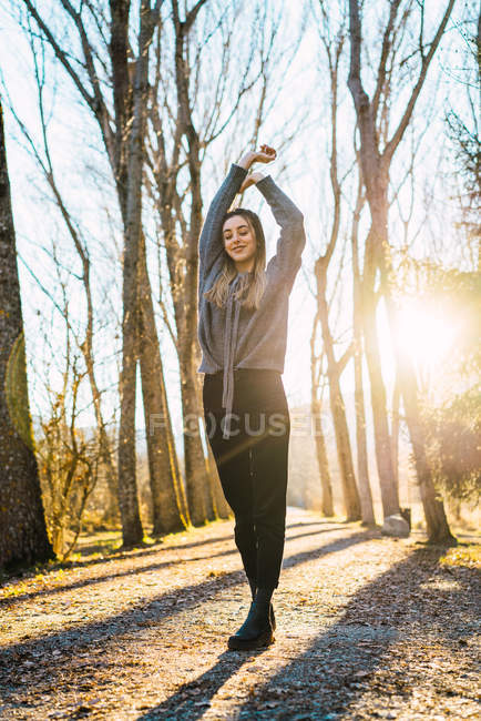 Bella donna con gli occhi chiusi posa con le braccia sollevate sul vicolo con file di alberi alla luce del sole
. — Foto stock