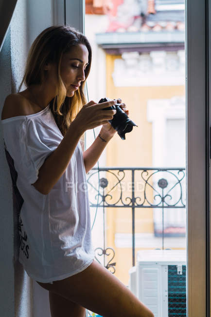Vista laterale del fotografo femminile che guarda scatti sulla fotocamera dalla finestra a casa — Foto stock