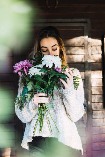 Mujer rubia en suéter de pie en el porche de la casa y oliendo ramo de flores - foto de stock