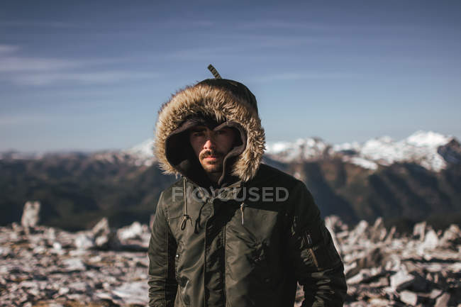 Porträt eines Mannes im warmen Mantel mit Kapuze, der vor dem Hintergrund schneebedeckter Berge posiert und in die Kamera blickt — Stockfoto