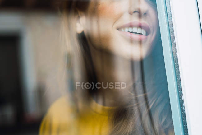 Вид через стекло улыбающейся женщины, стоящей и улыбающейся у окна дома . — стоковое фото