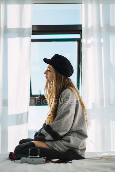 Vista laterale di giovane donna sognante in maglione e cappello seduta su letto bianco con fotocamera contro finestra . — Foto stock