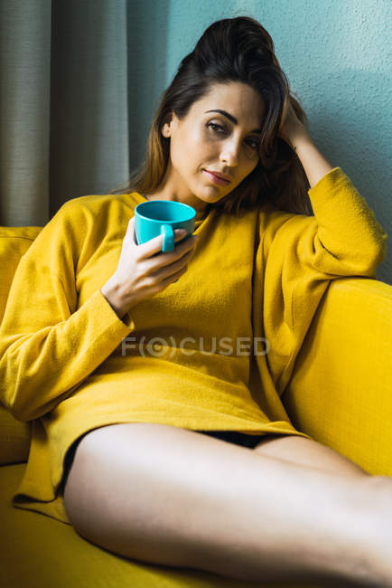 Nachdenkliche junge Frau mit einem Becher Heißgetränk am Kopf und entspannt im Sessel. — Stockfoto