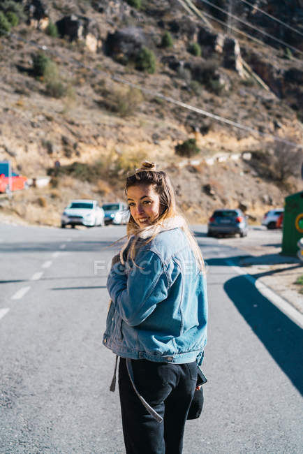 Back view of casual woman in denim jacket on roadway looking at camera over shoulder and smiling. — Stock Photo