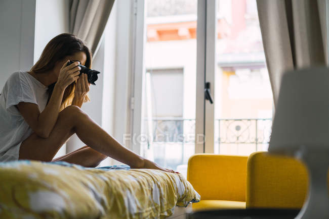 Vista laterale della donna che scatta con la fotocamera e si siede sul letto — Foto stock