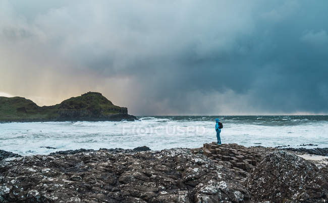 Отдаленный взгляд на туриста, стоящего на скалистом побережье и смотрящего на океан — стоковое фото
