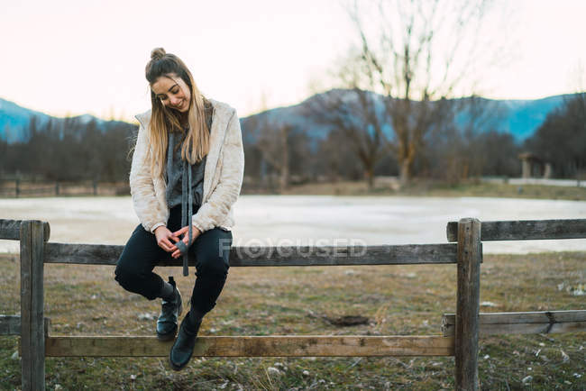 Portrait of woman in stylish warm clothing sitting on fence on rural background and smiling contently. — Stock Photo
