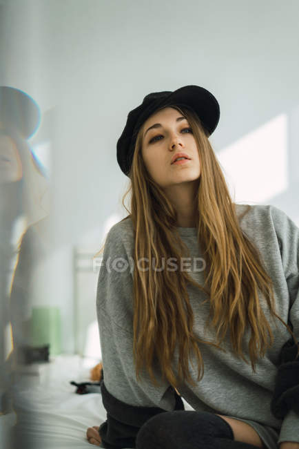 Porträt eines stilvollen jungen Mädchens mit Mütze, das auf dem Bett sitzt und wegschaut — Stockfoto