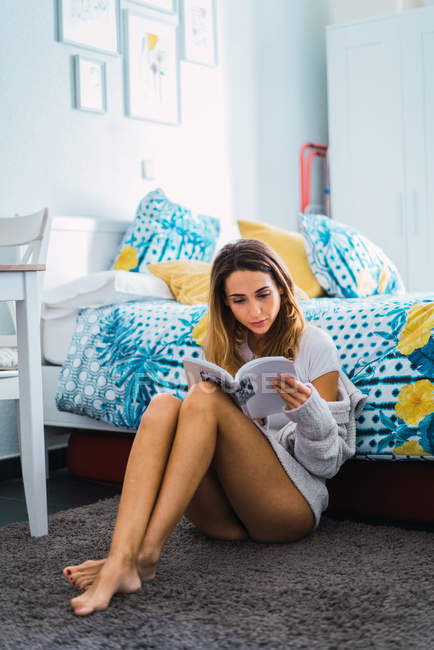 Retrato de la mujer sentada en el suelo por la cama y leyendo el libro - foto de stock