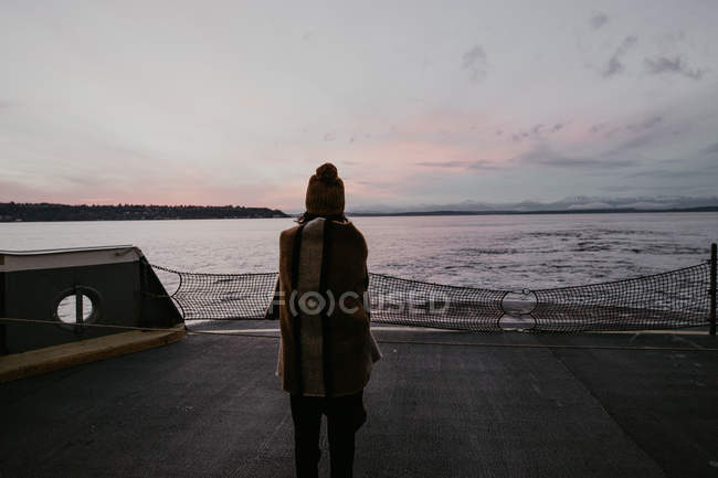 Vista trasera de la mujer envuelta en cuadros de pie en el barco y mirando hacia otro lado en el paisaje marino . - foto de stock