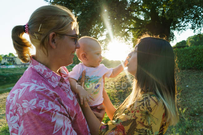 Vue latérale du couple lesbienne heureux avec enfant en bas âge dans un parc verdoyant ensoleillé . — Photo de stock