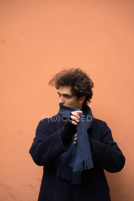 Retrato del joven que se pone la bufanda y mira hacia otro lado en el fondo de la pared naranja - foto de stock