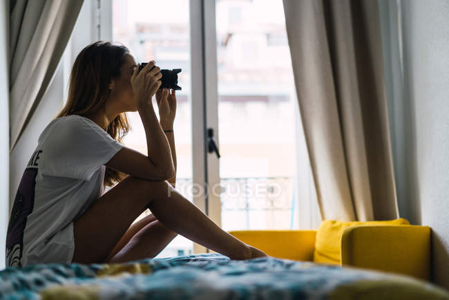 Vista lateral da jovem mulher bonita sentada na cama e focada com câmera em casa . — Fotografia de Stock