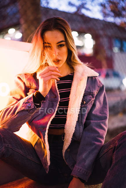 Portrait of woman in denim jacket looking at camera with provocation and sitting near lamplight on street — Stock Photo
