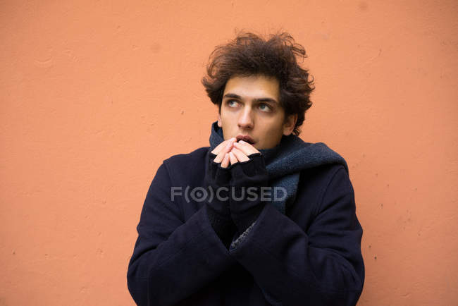 Retrato de homem jovem aquecendo as mãos com a respiração e olhando para cima no fundo da parede laranja — Fotografia de Stock