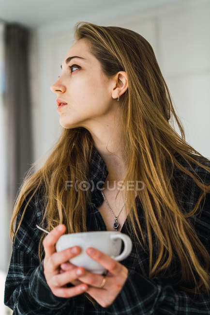 Portrait de fille aux cheveux longs tenant tasse de café à la maison et regardant ailleurs . — Photo de stock