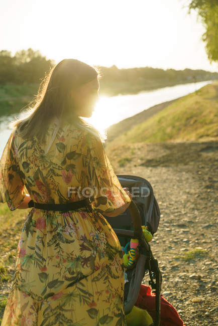 Rear view of young woman walking with baby carriage in park in sunlight — Stock Photo