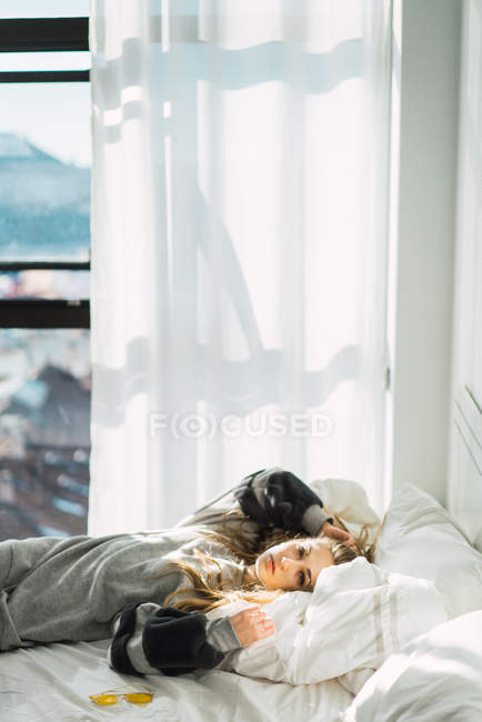 Blonde girl in gray sweatshirt lying on white sheet of bed looking at camera. — Stock Photo