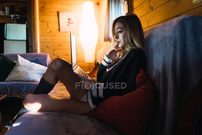 Vista lateral da mulher sensual em suéter assistindo laptop enquanto sentado no sofá em casa . — Fotografia de Stock