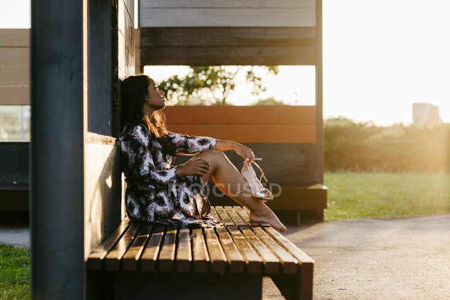 Side view of woman in dress sitting on bench with bare feet and holding shoes in hands — Stock Photo