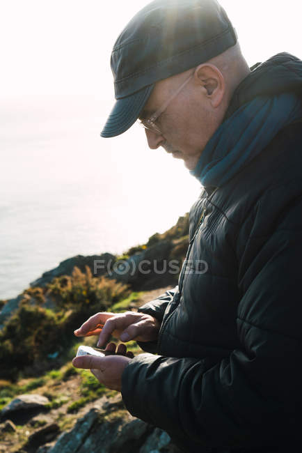 Vista lateral do homem em pé na colina verde e usando smartphone à beira-mar . — Fotografia de Stock