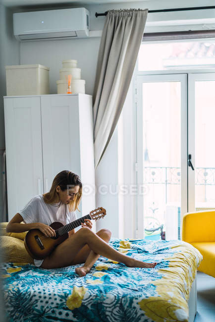 Side view of young woman playing guitar on bed at home — Stock Photo