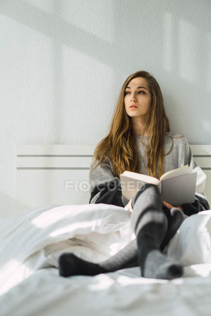 Chica relajante con libro en una cama acogedora y mirando hacia otro lado - foto de stock