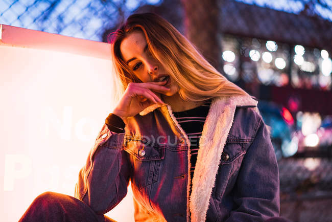 Portrait of woman in denim jacket looking at camera with provocation and sitting near lamplight on street in dusk — Stock Photo