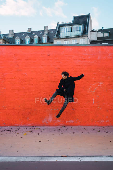 Vista lateral do homem em roupas quentes pulando na parede laranja na rua . — Fotografia de Stock