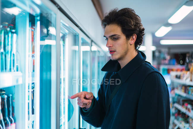Jeune homme en vêtements chauds debout au réfrigérateur en magasin et pointant du doigt — Photo de stock